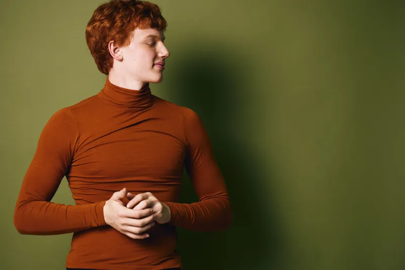 Young man with red hair wearing brown turtleneck sweater standing with closed eyes and calm expression on green background indoors.