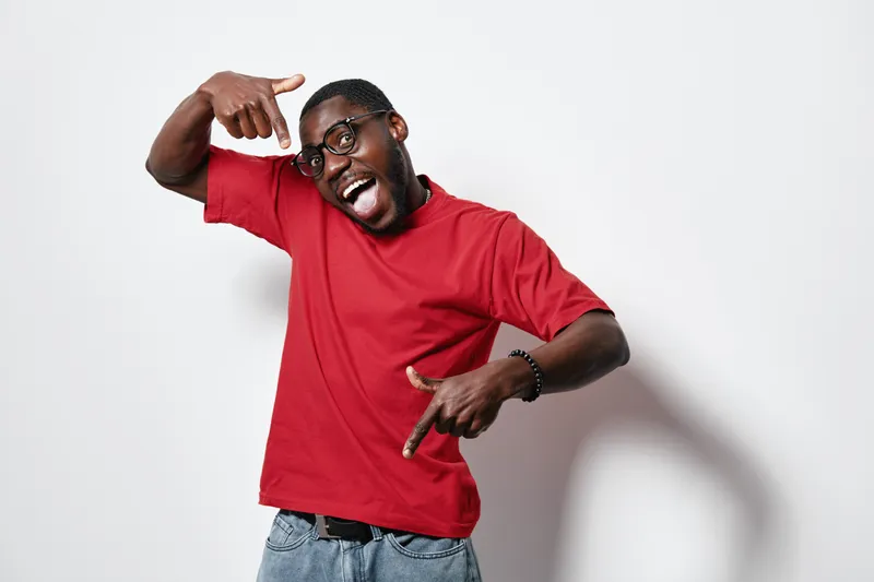 African American man with dark skin wearing red t shirt and glasses posing happily against white background with expressive gestures and joyful smile.