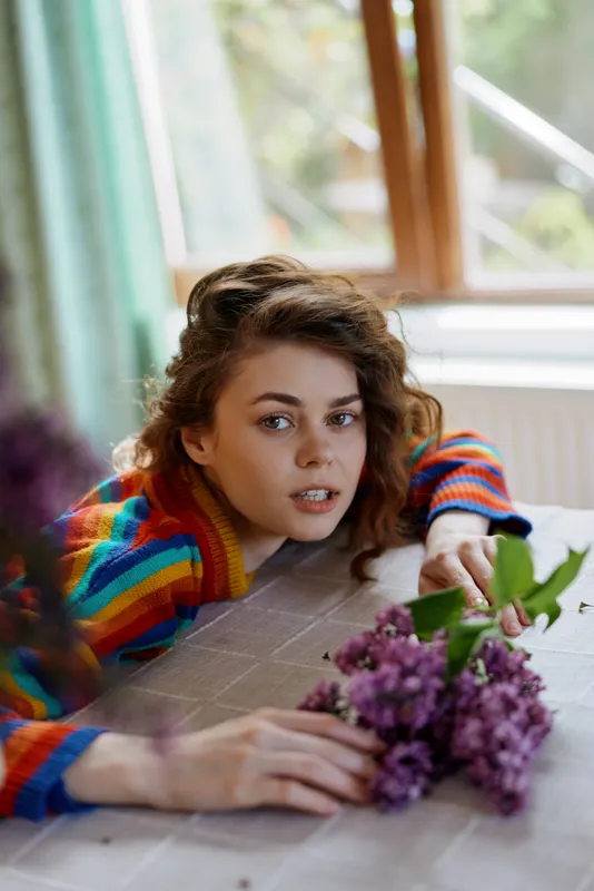 Cheerful woman indoors, lying at a table, wearing a colorful sweater, soft natural light, close to a bouquet, relaxed lifestyle moment in a cozy home setting with vibrant colors and calm mood