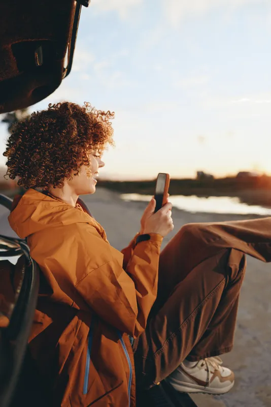 Woman lifestyle outdoors at sunset, curly hair and orange jacket with backpack, holding a smartphone and checking a phone screen, warm golden light, city street background, candid travel mood