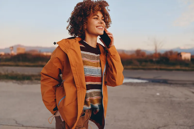 Woman lifestyle portrait outdoors at sunset by river, curly hair and orange jacket, smiling while holding phone to ear, casual travel mood, soft evening light, shallow depth, bokeh