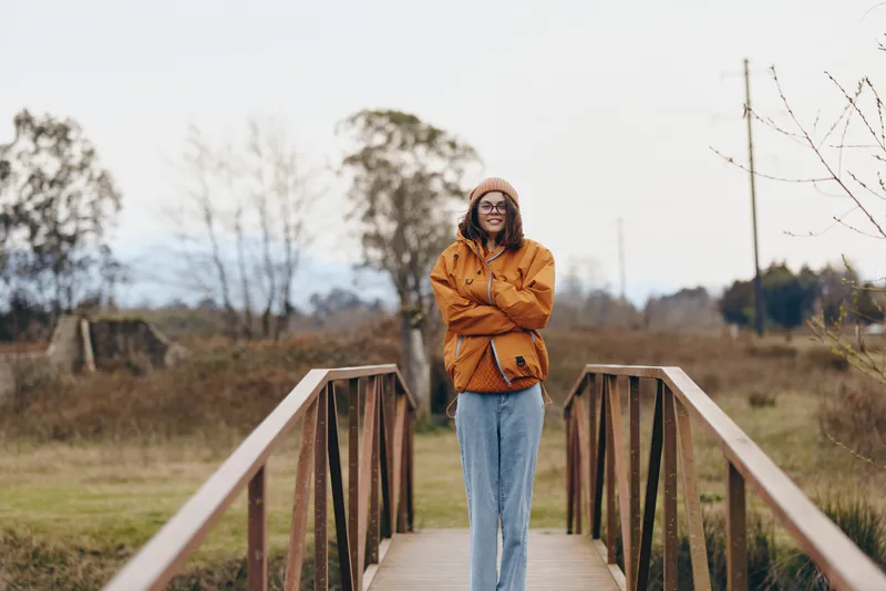 Woman lifestyle on a wooden bridge, standing outdoors in autumn park, wearing casual jacket and jeans, arms crossed, calm portrait at sunset, natural light, soft wide scenic background.