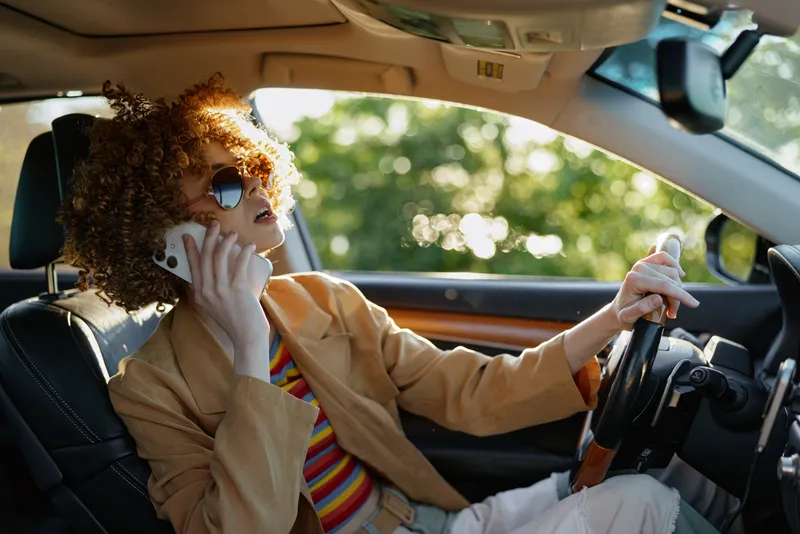 Young woman with curly hair rides in a car while talking on a cellphone, wearing a beige blazer and striped shirt in a retro film color vibe, the scene captures a candid, confident moment on the road