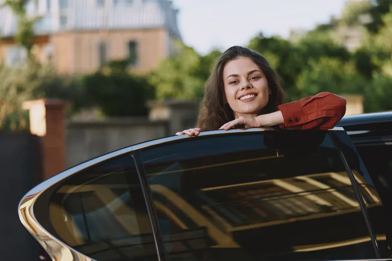 Cheerful woman outdoors, leaning from car window, smiling at camera, sunlit moment, relaxed posture, soft breeze, natural colors, lush greenery in background, carefree and confident vibe
