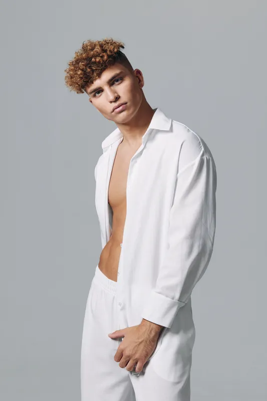 Young man with curly brown hair wearing white pants and an open oversized white shirt standing posing against a grey studio background looking at camera with serious expression.