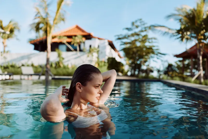 Woman relaxing in outdoor pool, adjusting hair, tropical resort background, sunny day, calm water surface, serene atmosphere, casual lifestyle moment