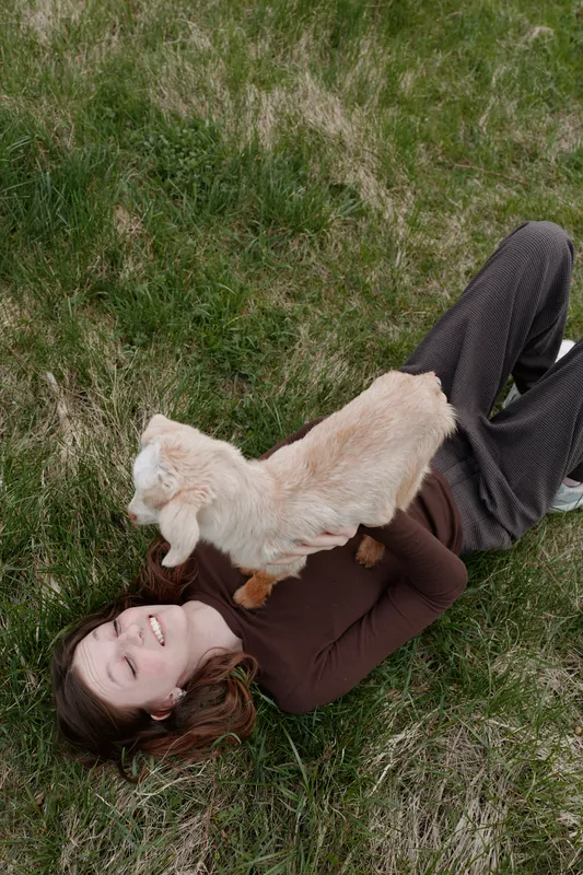 Lifestyle shot of a smiling woman lying on grass outdoors, goat kid climbing over her chest, affectionate moment, playful interaction, natural light, calm mood in a sunny park setting.