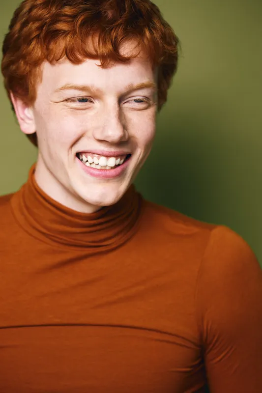 Young man with red hair wearing brown turtleneck smiling and looking sideways on green background. Portrait of happy male with freckles and casual fashion style in studio shot.