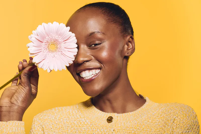 Happy African woman with dark skin smiles holding pink flower near face on yellow background. Concept of beauty, skincare, natural glow, and positive mood in studio portrait.