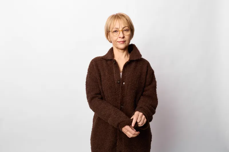 Woman portrait studio, senior woman in brown coat and glasses, calm smile, standing indoors against plain white background, thoughtful relaxed mood, soft lighting