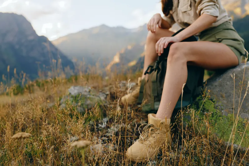 Young woman resting on a hillside trail, legs crossed, wearing combat boots and olive shorts, enjoying a quiet outdoor moment and the warm late sun.