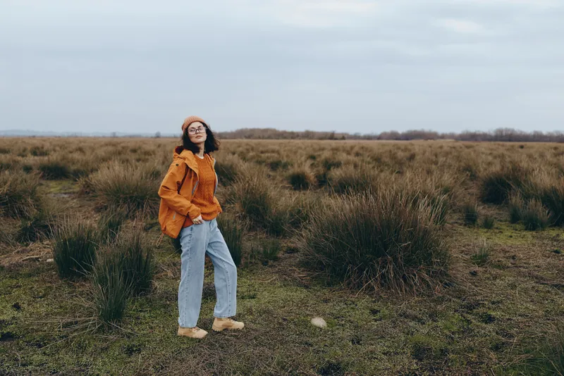 Woman, casual lifestyle, outdoors, grassy field, wearing orange sweater and light jeans, standing in the meadow, looking at camera, cloudy sky, calm natural light, lifestyle portrait, wind