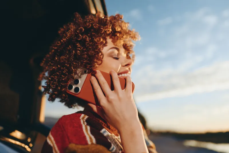 Woman lifestyle portrait outdoors at sunset, curly hair woman holding smartphone to ear, making phone call, relaxed smile, warm evening light, street background, casual daily life