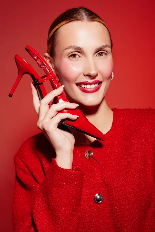Smiling woman in red sweater holds red high heel shoe near face on red background. Concept of fashion, style, beauty, and confident female model with bright makeup and accessories.