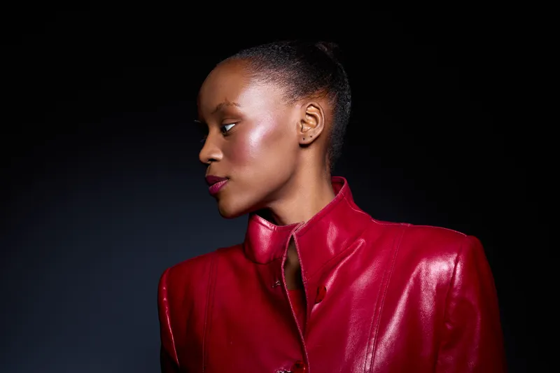 Profile of an elegant black woman with dark skin wearing a shiny red leather jacket, looking sideways against a dark background. Concept of fashion, beauty, and confidence.