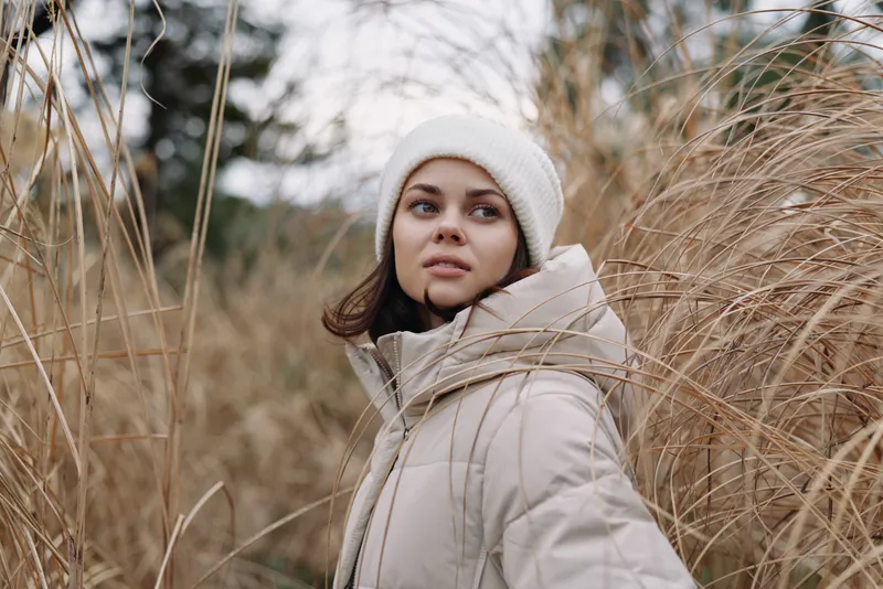 Woman in a beige winter coat and beanie stands among tall dried grasses in an outdoor setting, capturing a calm, candid portrait with soft natural light.