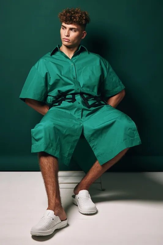 Young man with curly hair wearing green oversized shirt and shorts sitting on stool against dark background with white slip on shoes and casual pose.