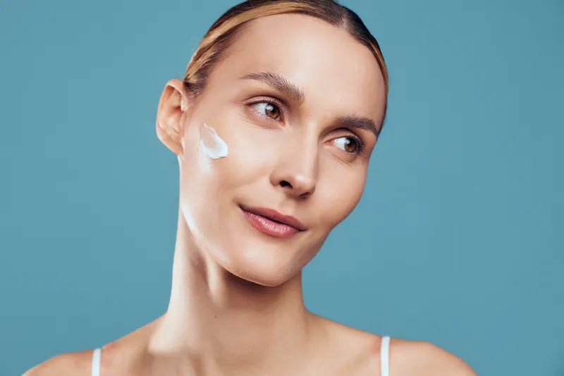 Young woman with clear skin applying white face cream on cheek, looking away. Blue background, natural makeup, skincare routine, wellness and beauty concept. Soft lighting.