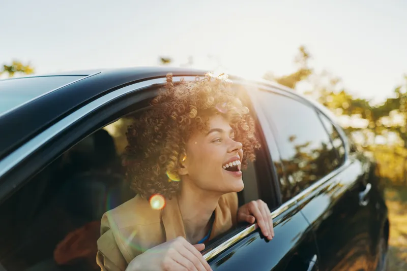 Smiling woman with curly hair leaning out of a car window, sunlight on a sunny day, retro mood, candid lifestyle moment, warm and carefree.