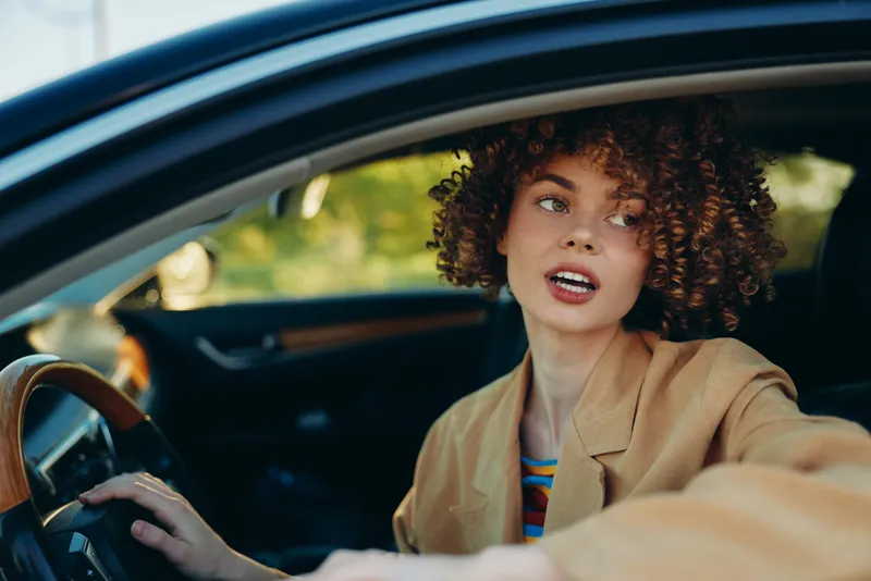 Cheerful woman driving car, looking aside, sunlit interior moment, casual jacket, warm natural colors, candid lifestyle scene inside vehicle outdoor glow, easygoing pose, sunshine, relaxed