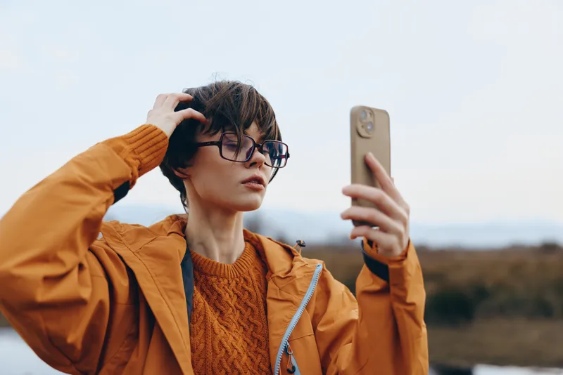 Young woman in casual orange jacket takes selfie with smartphone outdoors, adjusts hair, wears glasses, calm lifestyle portrait, natural light, blurred landscape background, technology moment