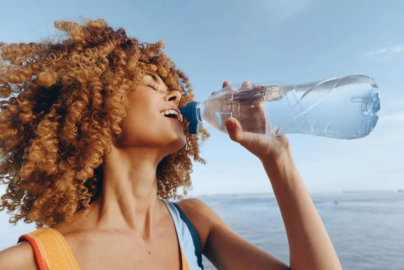 Curly woman drinking water from a plastic bottle outdoors on a sunny day. Blue sky and sea background. Concept of hydration, health, and summer lifestyle.