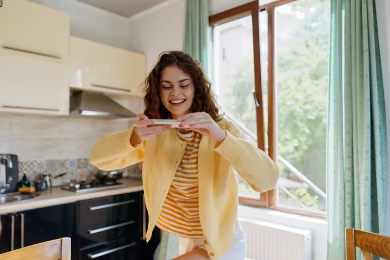 A cheerful woman in a kitchen checks her phone, a casual home scene with morning light streaming through a window, portraying everyday connection and warmth.