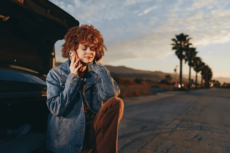 Woman with curly hair in denim jacket, holding a phone near open car at sunset, outdoor lifestyle concept, casual everyday moment, warm light on her face, desert road background nearby