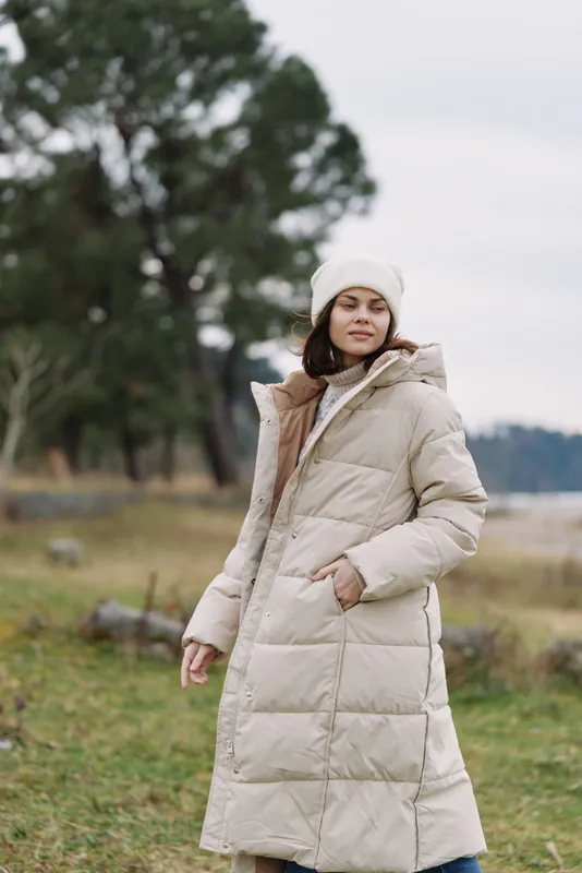 Woman in a beige winter coat stands outdoors in a calm field, showcasing fashion and portrait style in a serene countryside scene with a chilly cozy vibe.