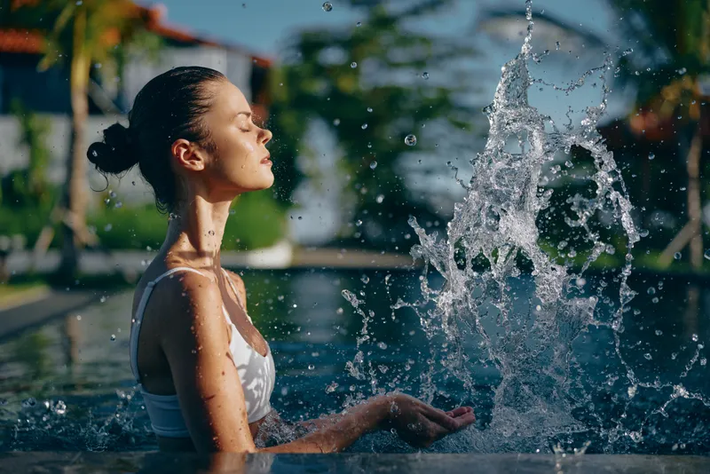 Young woman in white swimsuit enjoying water splash in outdoor pool. Relaxed lifestyle scene with natural sunlight and tropical background. Concept of wellness and summer leisure.