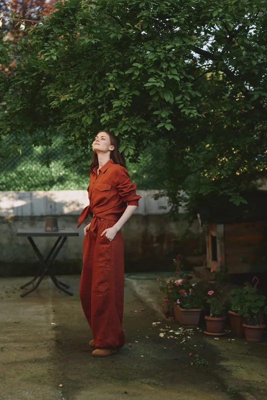 Woman in a rust red jumpsuit gazes upward in a garden, surrounded by greenery and potted plants. Casual, serene lifestyle scene with natural light and warm mood