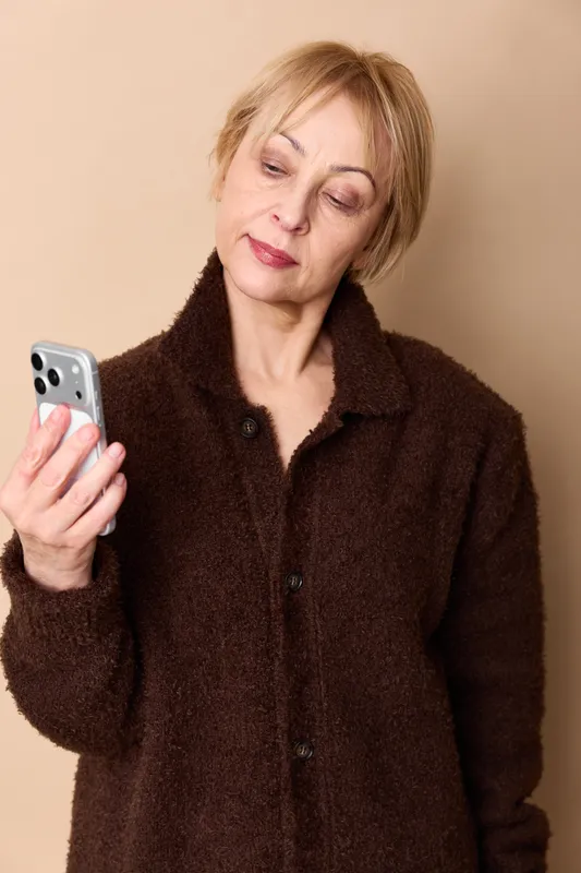 Woman smartphone elderly stress anxiety pain phone portrait, eyes closed expression, holding mobile in hand, dark fur coat, neutral beige indoor background