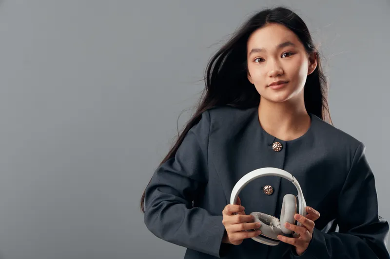 Woman with long dark hair holding white headphones wearing a dark jacket standing against grey background looking at camera with calm expression and relaxed posture.
