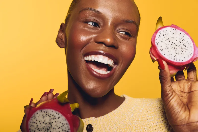 Happy African woman with dark skin holding two halves of dragon fruit on yellow background. Close-up portrait with bright lighting and joyful expression. Concept of healthy lifestyle and freshness.