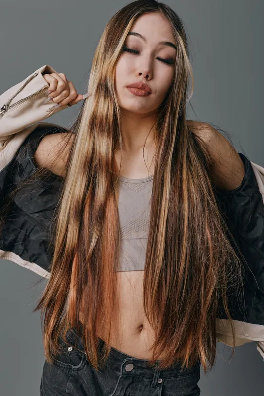 Young woman with long straight hair wearing a crop top and jacket posing against gray background with closed eyes, showing fashion and casual style in studio portrait.