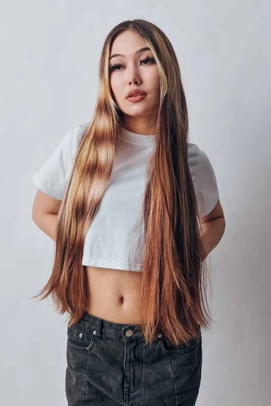 Young woman with long straight hair wearing white cropped t shirt and black jeans standing against plain white background with serious facial expression.