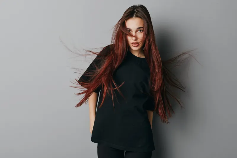Young woman with long red hair wearing black t shirt standing against gray background looking at camera with confident expression and natural makeup in studio portrait.
