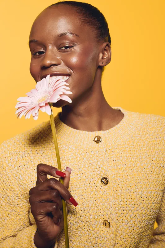 Smiling dark skinned African woman holding pink flower near face wears yellow textured sweater on bright yellow background. Concept of beauty, happiness, skincare, and natural glow.