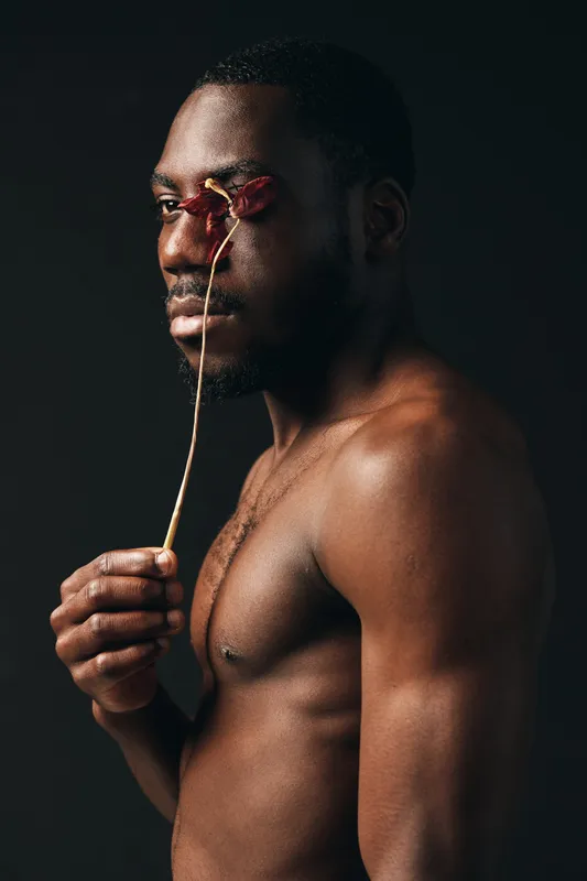 Creative conceptual portrait of African man with bare torso holding dried flower over eyes in dark studio. Artistic visual style with focus on skin texture and emotional expression.