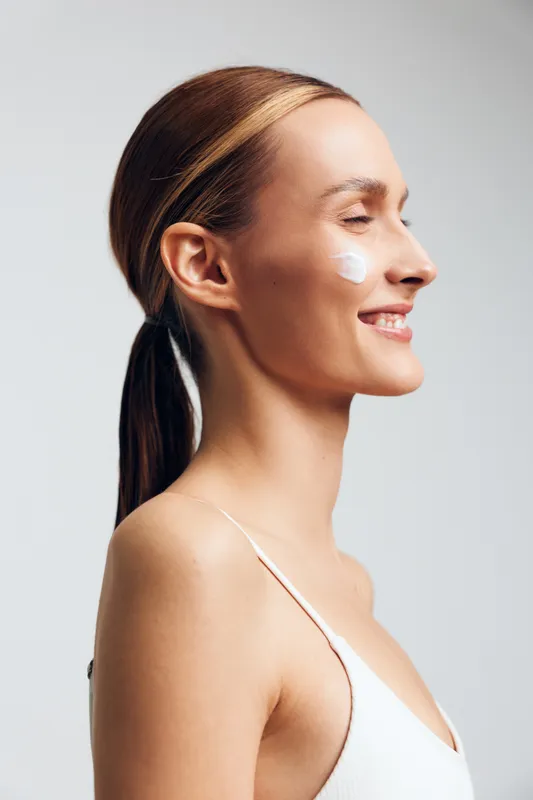 Smiling woman in white top applying cream on cheek, side profile, healthy skin, natural beauty, skincare routine, clean background, soft lighting