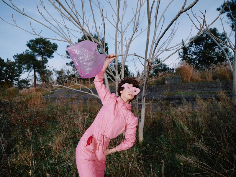 Dancer in a pink jumpsuit swirls a scarf beside a leafless tree in a grassy field, capturing expressive motion, theatrical style, and a dreamlike outdoor fashion moment