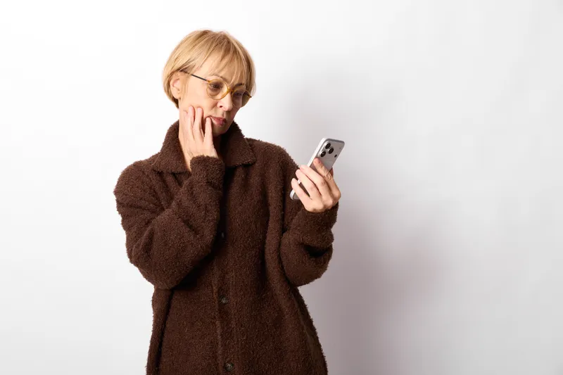 Woman holding smartphone, surprised worried expression, hand near mouth, glasses and coat in white studio setting, minimal background, soft light, clean copyspace area for text