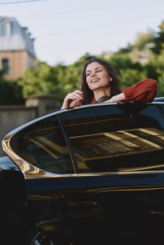 Cheerful woman leaning from car window, smiling outdoors, sunlit moment, relaxed pose, fresh air, casual fashion, natural colors, lush greenery background with soft breeze