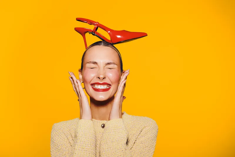Happy woman with closed eyes smiles broadly wearing red lipstick and beige sweater. Bright orange background and red high heel shoes on her head create playful mood.