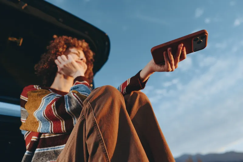 Woman holding smartphone outdoors near open car trunk, travel lifestyle under blue sky, casual jacket and sweater, she looks at the screen in daylight