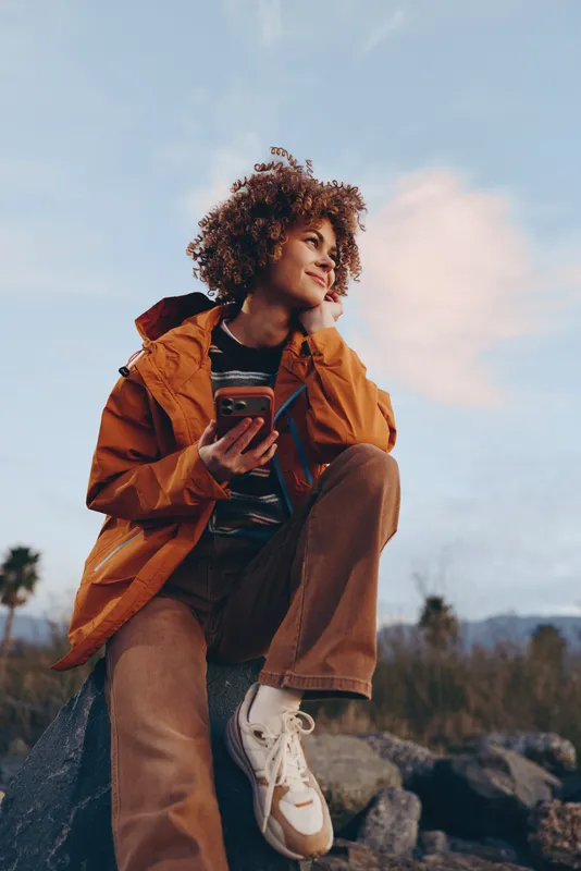 Woman lifestyle portrait outdoors, sitting on rocks, wearing orange jacket, curly hair, looking at smartphone, golden sky with soft clouds, calm relaxed travel mood, nature background