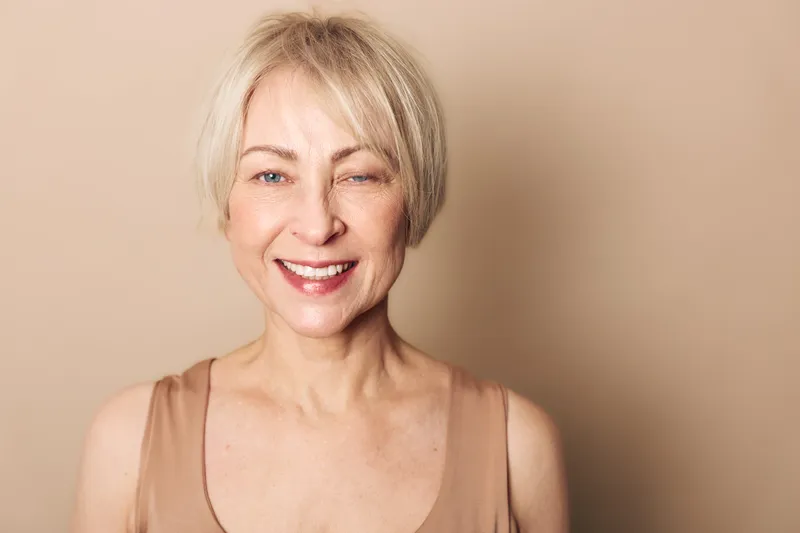 Happy mature woman with short blonde hair and natural makeup smiles gently against beige background. Portrait of confident middle aged female with positive expression and casual style.