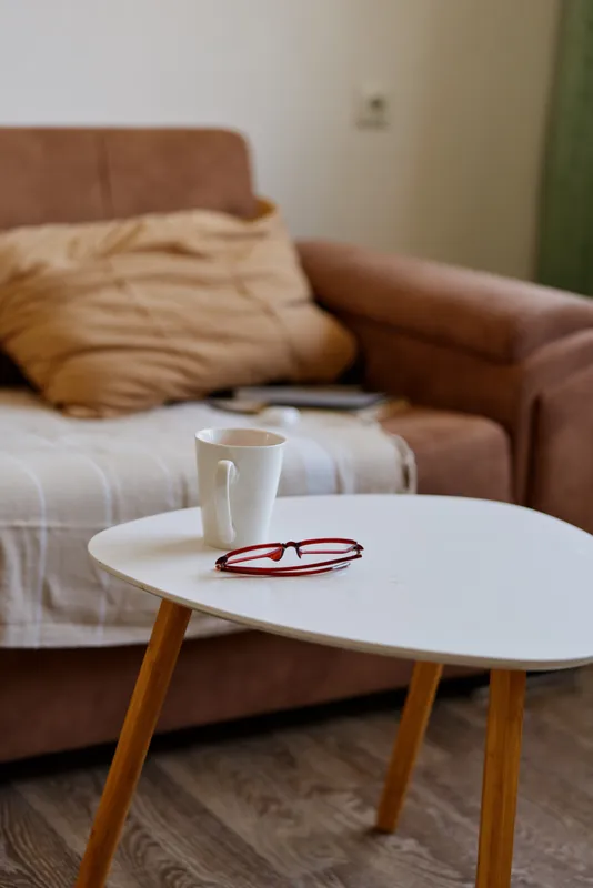 Cozy living room scene with a white round table, a mug, red glasses, a soft blanket on a brown sofa, warm natural light, calm domestic atmosphere and relaxed home mood.