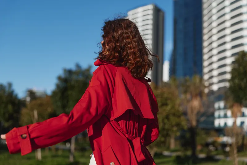 Young woman with curly hair wearing a red coat spins outdoors in an urban park with tall buildings in the background on a sunny day.