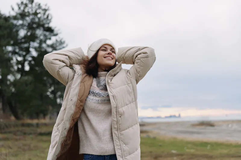 Outdoor woman wearing a beige jacket and sweater, standing in an open landscape with trees, smiling and relaxing with hands behind head